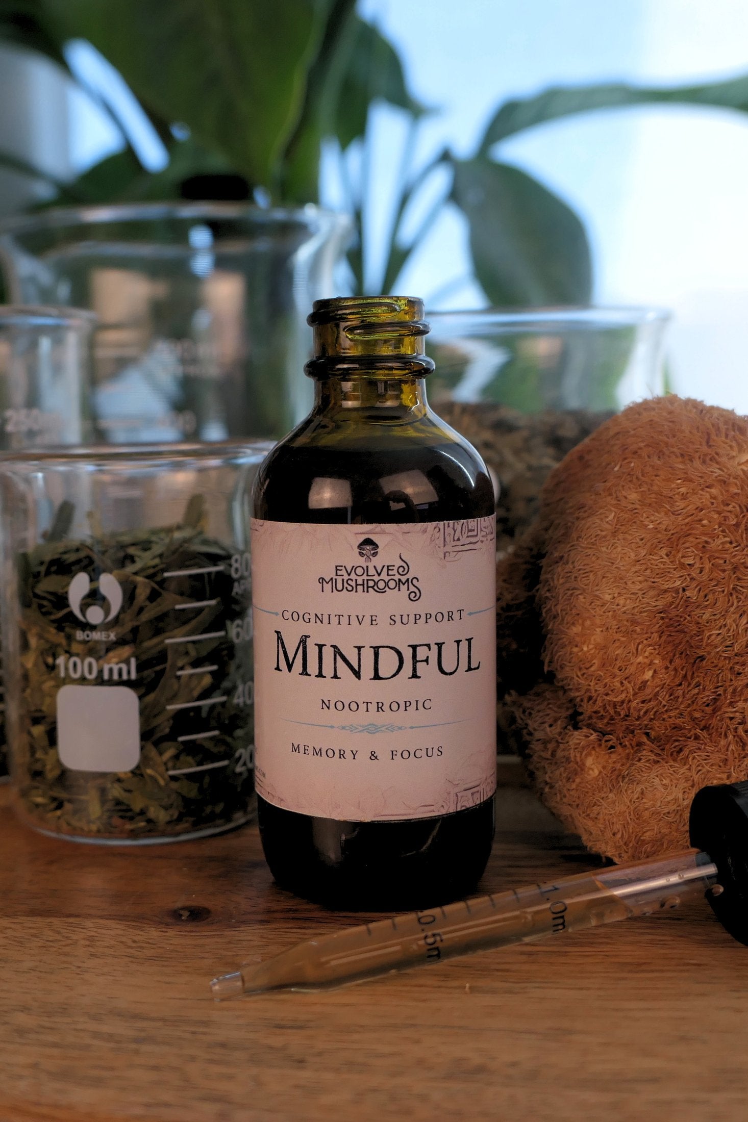 Angled shot of Evolved Mushrooms Mindful nootropic dropper bottle with a glass dropper tool, staged with lab beakers and dried herbs on a wood surface.